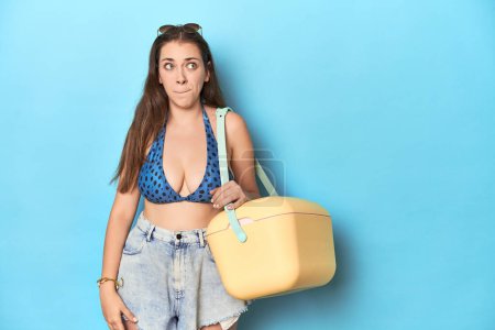Photo for Woman in bikini with a portable beach cooler, blue studio confused, feels doubtful and unsure. - Royalty Free Image