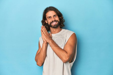 Photo for Bearded man in a tank top, blue backdrop feeling energetic and comfortable, rubbing hands confident. - Royalty Free Image