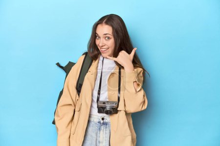 Photo for Young Caucasian woman traveler with camera and backpack showing a mobile phone call gesture with fingers. - Royalty Free Image