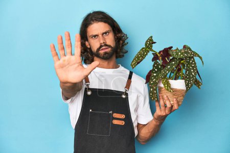 Téléchargez les photos : Jardinier aux cheveux longs avec barbe tenant une plante debout avec la main tendue montrant le panneau d'arrêt, vous empêchant. - en image libre de droit