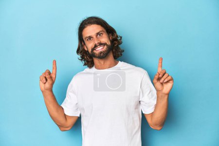 Photo for Bearded man in a white shirt, blue backdrop indicates with both fore fingers up showing a blank space. - Royalty Free Image
