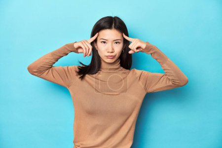 Photo for Young Asian woman in brown turtleneck, focused on a task, keeping forefingers pointing head. - Royalty Free Image
