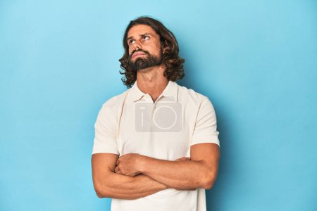 Photo for Long-haired man in a white polo, blue studio tired of a repetitive task. - Royalty Free Image