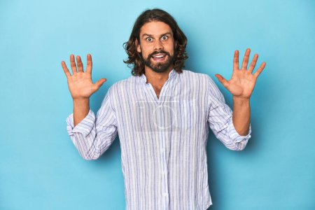 Photo for Man with beard in blue striped shirt, blue studio receiving a pleasant surprise, excited and raising hands. - Royalty Free Image