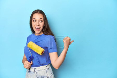 Photo for Young woman with yellow paint roller on a blue background points with thumb finger away, laughing and carefree. - Royalty Free Image