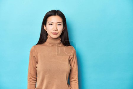 Photo for Asian woman in brown turtleneck looking at camera, blue background. - Royalty Free Image
