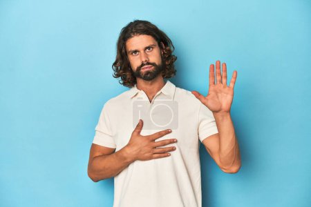 Photo for Long-haired man in a white polo, blue studio taking an oath, putting hand on chest. - Royalty Free Image