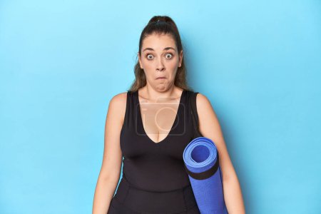 Photo for Young sporty Caucasian woman with yoga mat in blue studio Young sporty Caucasian woman with yoga mat in blue studioshrugs shoulders and open eyes confused. - Royalty Free Image