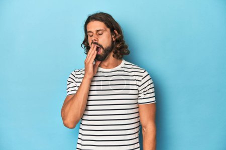 Foto de Hombre barbudo con camisa a rayas, fondo azul bostezando mostrando un gesto cansado cubriendo la boca con la mano. - Imagen libre de derechos