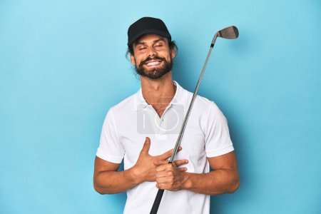 Foto de Golfista de pelo largo con club y sombrero se ríe en voz alta manteniendo la mano en el pecho. - Imagen libre de derechos