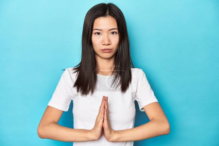 Foto de Mujer asiática joven en camiseta blanca, plano de estudio, rezando, mostrando devoción, persona religiosa en busca de inspiración divina. - Imagen libre de derechos
