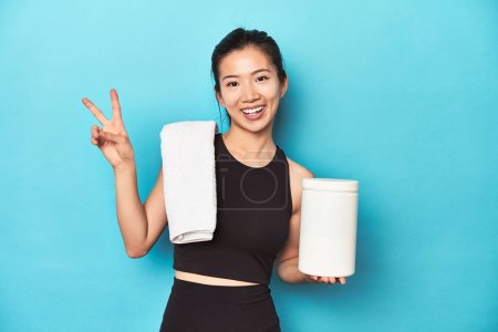 Photo for Young Asian sportswoman with protein bottle, gym setup, joyful and carefree showing a peace symbol with fingers. - Royalty Free Image