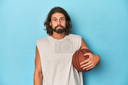 Photo for Bearded man with basketball in blue studio shrugs shoulders and open eyes confused. - Royalty Free Image