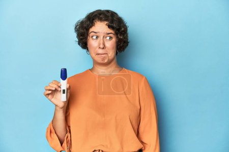 Photo for Young woman holding pregnancy test, studio background confused, feels doubtful and unsure. - Royalty Free Image