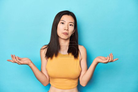 Photo for Asian woman in summer yellow top, studio setup, doubting and shrugging shoulders in questioning gesture. - Royalty Free Image