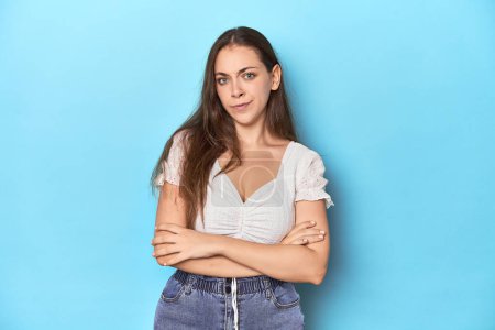 Photo for Stylish young woman in white blouse on a blue studio backdrop suspicious, uncertain, examining you. - Royalty Free Image