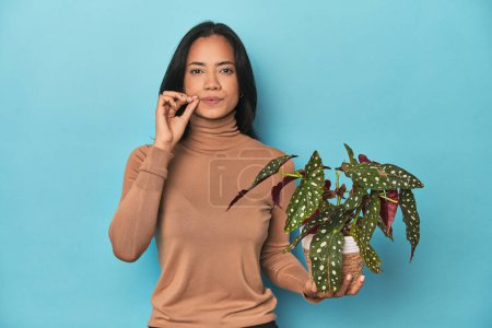 Téléchargez les photos : Filipina tenant une plante sur le studio bleu avec les doigts sur les lèvres gardant un secret. - en image libre de droit
