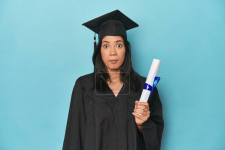 Photo for Filipina graduate with diploma on blue studio shrugs shoulders and open eyes confused. - Royalty Free Image