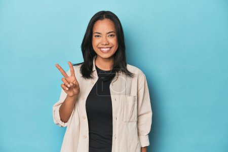 Photo for Filipina young woman on blue studio showing victory sign and smiling broadly. - Royalty Free Image
