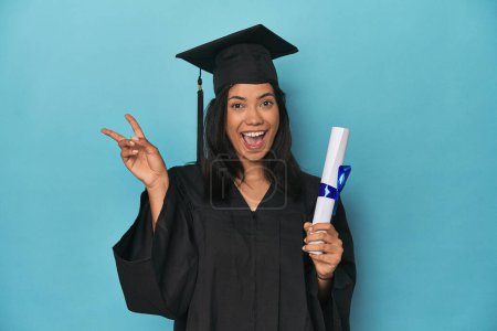 Téléchargez les photos : Filipina diplômé avec diplôme sur studio bleu joyeux et insouciant montrant un symbole de paix avec les doigts. - en image libre de droit