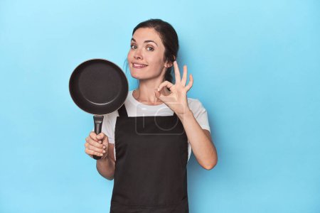 Photo for Woman with apron and pan on blue background cheerful and confident showing ok gesture. - Royalty Free Image