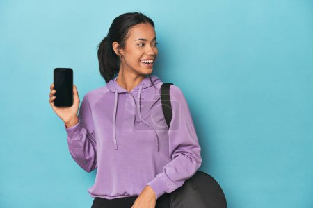 Photo for Filipina ready for gym with phone on blue looks aside smiling, cheerful and pleasant. - Royalty Free Image