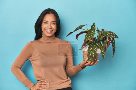 Foto de Filipina sosteniendo una planta en el estudio azul mostrando un espacio de copia en una palma y sosteniendo otra mano en la cintura. - Imagen libre de derechos