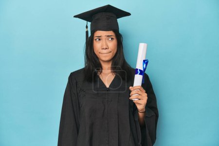 Photo for Filipina graduate with diploma on blue studio confused, feels doubtful and unsure. - Royalty Free Image