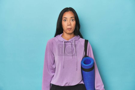 Photo for Filipina athlete with yoga mat on blue shrugs shoulders and open eyes confused. - Royalty Free Image