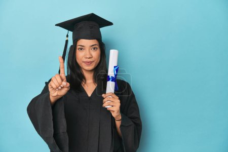Photo for Filipina graduate with diploma on blue studio showing number one with finger. - Royalty Free Image