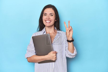Téléchargez les photos : Jeune femme d'affaires tenant un ordinateur portable sur bleu montrant le numéro deux avec les doigts. - en image libre de droit