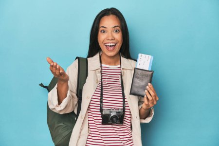Téléchargez les photos : Filipina avec caméra, billets, sac à dos sur bleu recevant une agréable surprise, excité et levant la main. - en image libre de droit