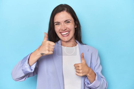 Woman in blue blazer on blue background raising both thumbs up, smiling and confident.