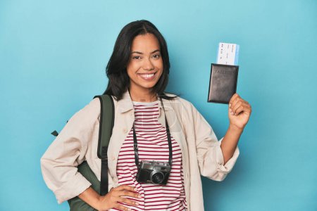Téléchargez les photos : Filipina avec appareil photo, billets, sac à dos sur bleu montrant un espace de copie sur une paume et tenant une autre main à la taille. - en image libre de droit