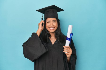 Foto de Filipina graduada con diploma en estudio azul cubriendo orejas con las manos. - Imagen libre de derechos