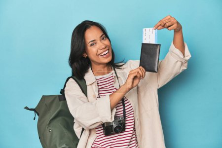 Photo for Filipina traveler displaying flight ticket with camera and backpack on blue - Royalty Free Image