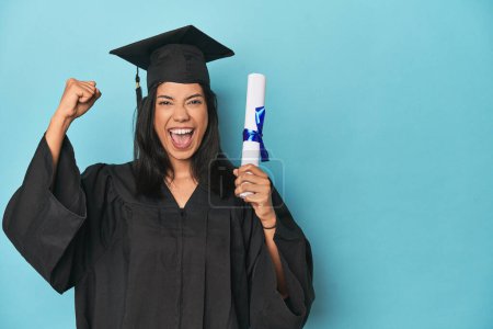 Foto de Filipina se gradúa con diploma en estudio azul levantando puño después de una victoria, concepto ganador. - Imagen libre de derechos