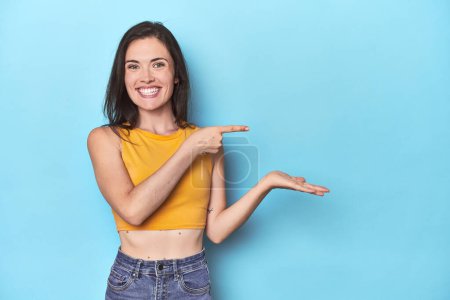 Photo for Young caucasian woman on blue backdrop excited holding a copy space on palm. - Royalty Free Image