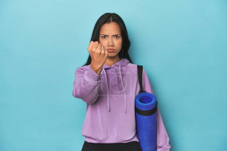 Foto de Atleta filipina con esterilla de yoga en azul mostrando puño a cámara, expresión facial agresiva. - Imagen libre de derechos