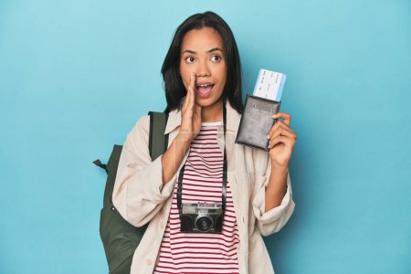 Photo for Filipina with camera, tickets, backpack on blue is saying a secret hot braking news and looking aside - Royalty Free Image