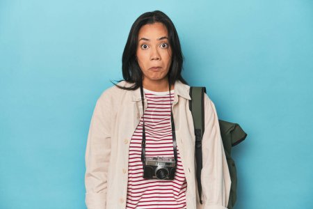 Photo for Filipina woman with camera and backpack on blue shrugs shoulders and open eyes confused. - Royalty Free Image