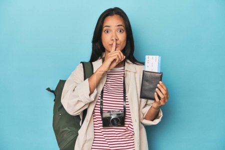 Photo for Filipina with camera, tickets, backpack on blue keeping a secret or asking for silence. - Royalty Free Image