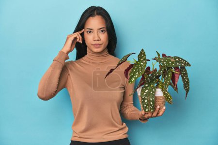 Photo for Filipina holding a plant on blue studio pointing temple with finger, thinking, focused on a task. - Royalty Free Image