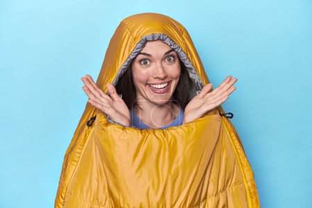 Photo for Woman in sleeping bag on blue background receiving a pleasant surprise, excited and raising hands. - Royalty Free Image