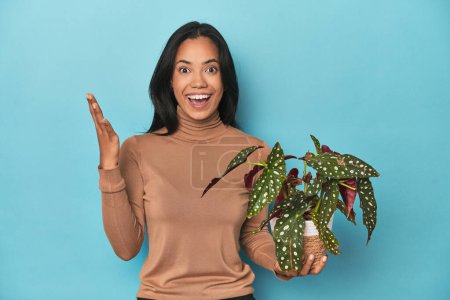 Photo for Filipina holding a plant on blue studio receiving a pleasant surprise, excited and raising hands. - Royalty Free Image