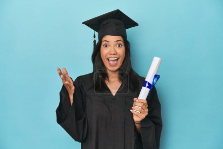Foto de Filipina se gradúa con diploma en estudio azul recibiendo una agradable sorpresa, emocionado y levantando las manos. - Imagen libre de derechos