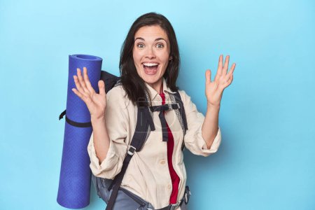 Foto de Mujer aventurera con mochila en azul recibiendo una agradable sorpresa, excitada y levantando las manos. - Imagen libre de derechos