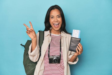Photo for Filipina with camera, tickets, backpack on blue joyful and carefree showing a peace symbol with fingers. - Royalty Free Image