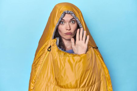 Photo for Woman in sleeping bag on blue background standing with outstretched hand showing stop sign, preventing you. - Royalty Free Image