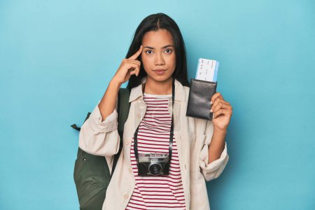 Photo for Filipina with camera, tickets, backpack on blue pointing temple with finger, thinking, focused on a task. - Royalty Free Image